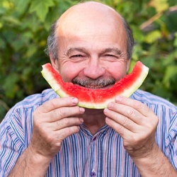 An older man holds up a piece of watermelon and smiles