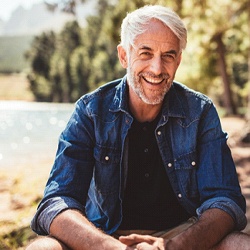 An older man sitting outside by the water and smiling after meeting with his dentist to discuss the potential for full-mouth reconstruction